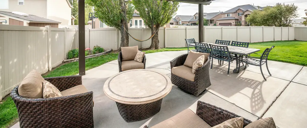 A spacious backyard concrete patio area furnished with woven wicker seating and a round table, overlooking a fenced green lawn and a hammock between two trees.