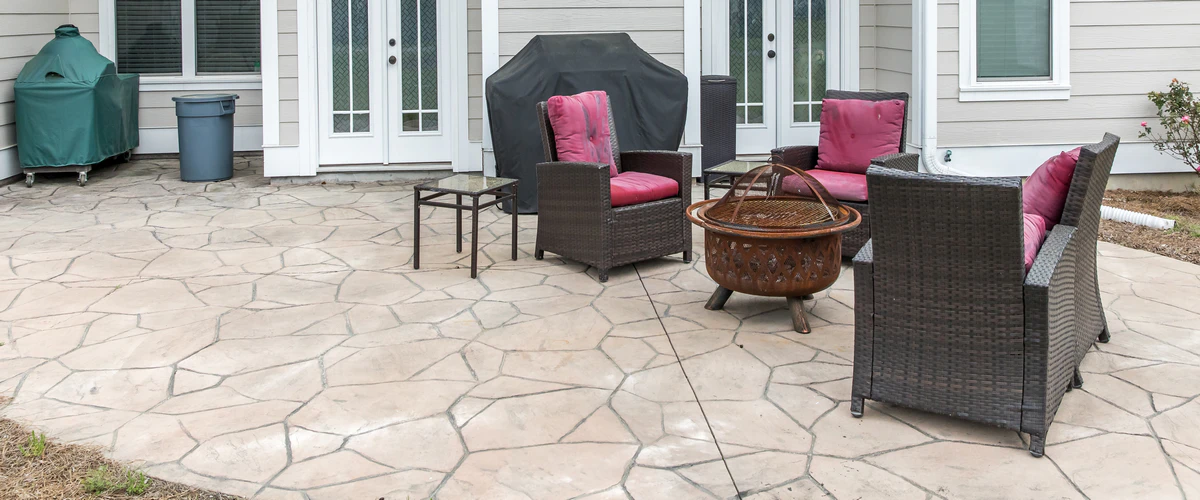 A spacious backyard patio area featuring light-colored flagstone style concrete staining, complete with outdoor patio chairs, a rusty fire pit, and a covered grill.