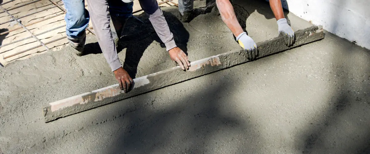 Close-up of two contractors working together to pull a long straight edge board across wet cement for precise concrete leveling.